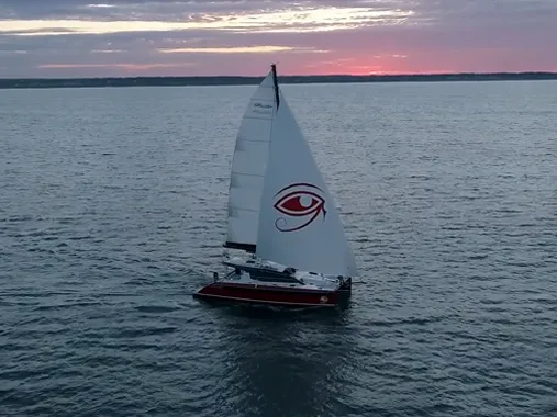 Sailboat with eye-like logo navigates calm waters during a vibrant sunset, capturing serene nautical beauty.