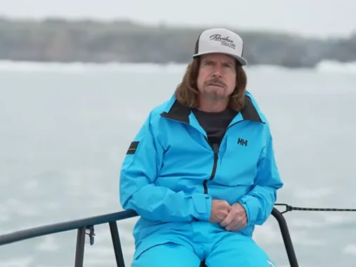 Man in blue sailing jacket and cap sitting on a boat against a calm water backdrop.